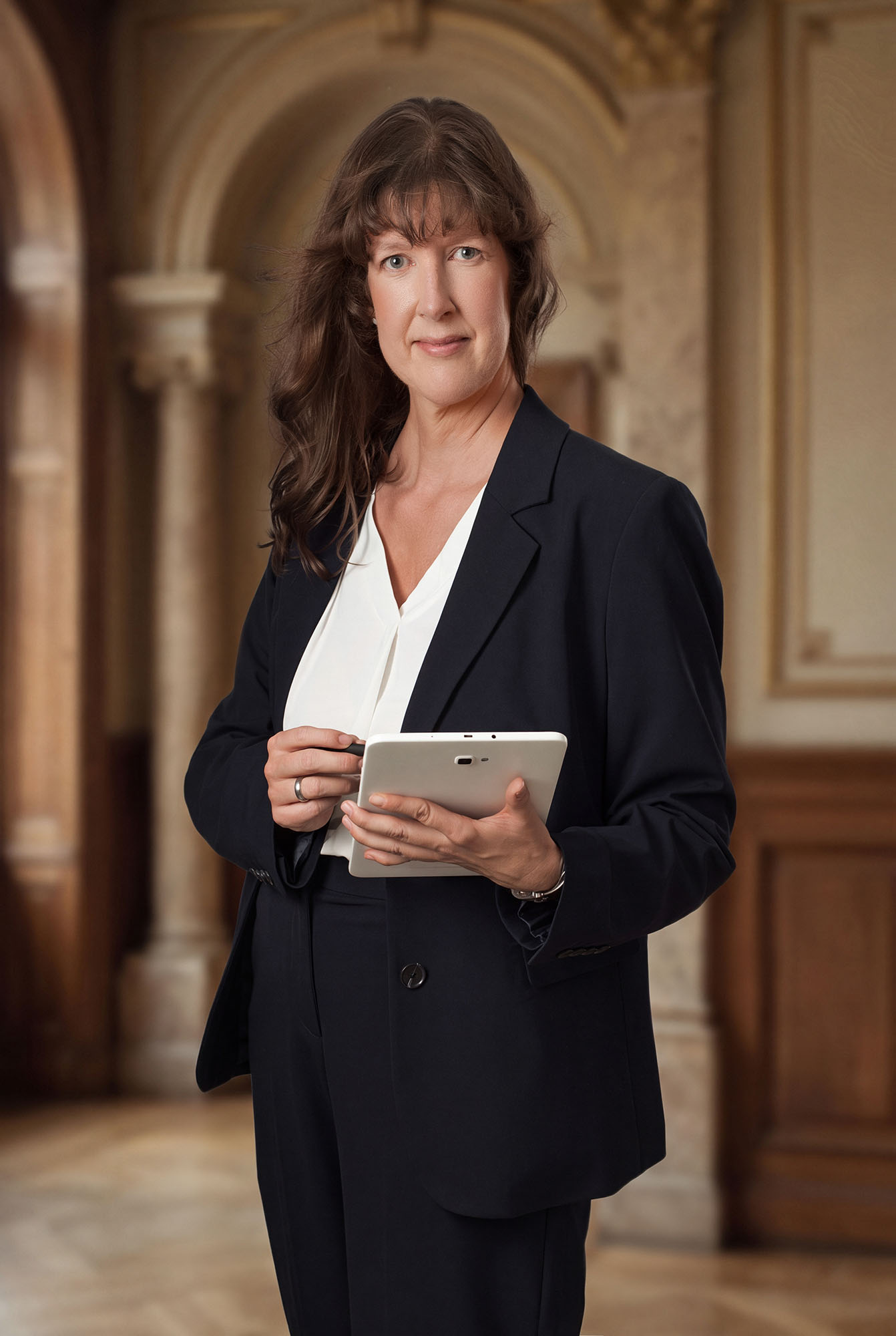 Businessfotografie Personal Branding Pfaffenhofen: Anwältin im dunklen Anzug hält Tablet in klassischer Eingangshalle, Anton Gil Fotografie.